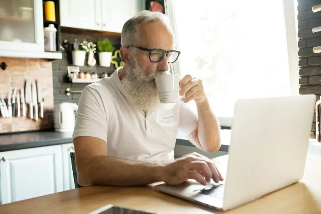 Older man working on laptop, smiling, looking at screen, drinking tea.